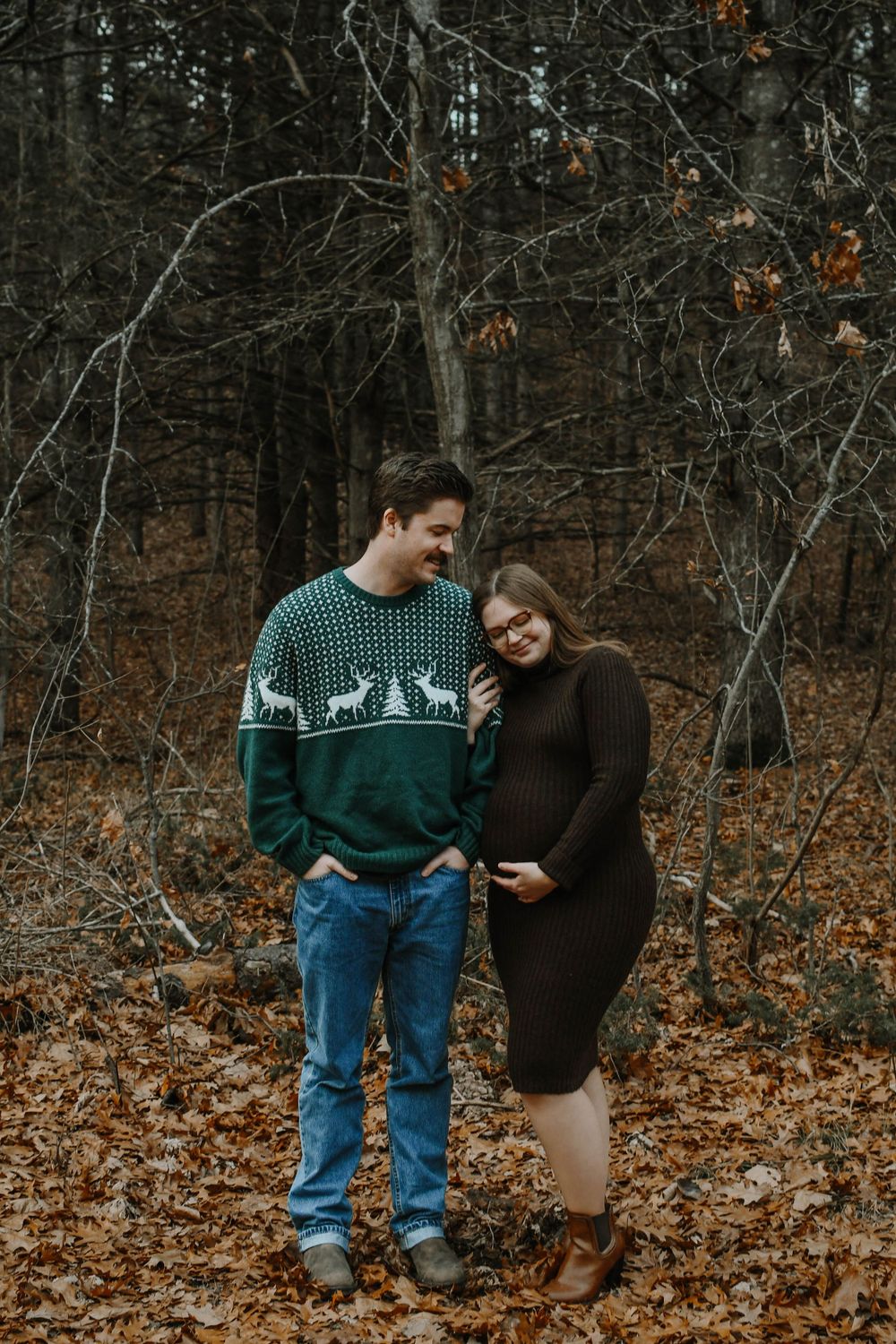 A couple stands close together on fallen autumn leaves in the woods, one wearing a festive reindeer sweater.