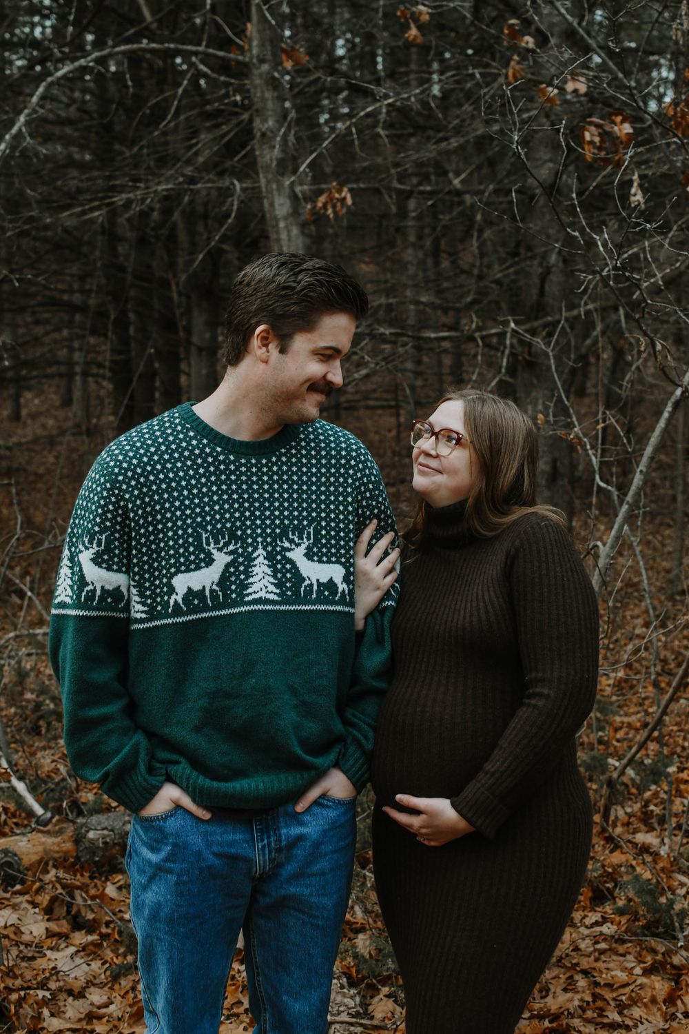 Couple in autumn woods sharing intimate moment with festive holiday sweater and brown knit dress during maternity photoshoot.