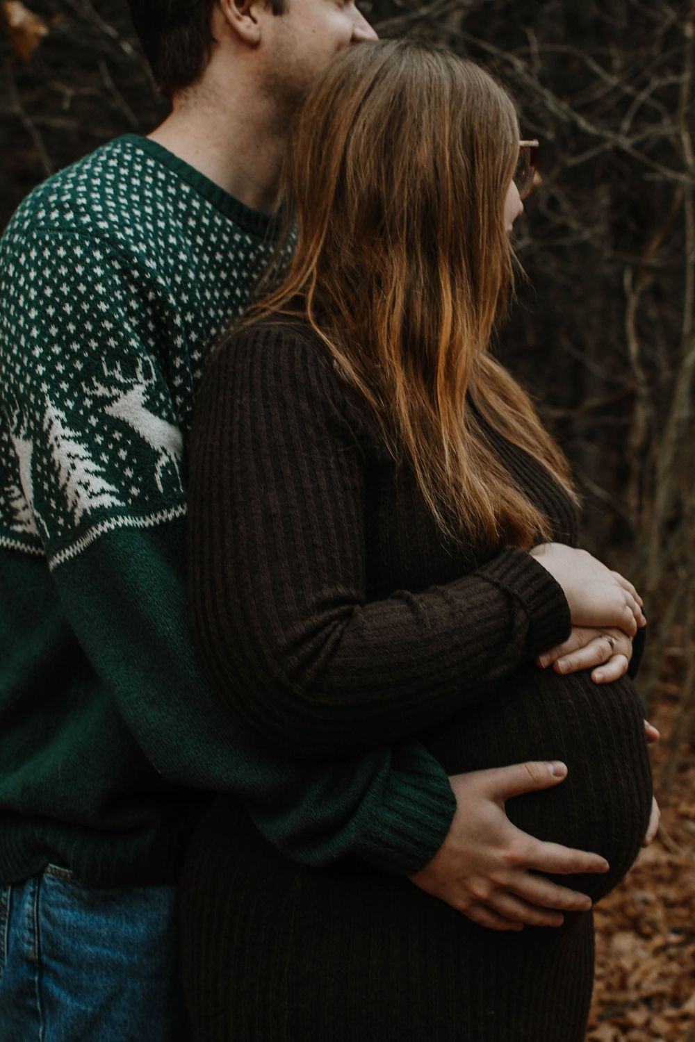 A pregnant woman in a black sweater being embraced from behind by someone wearing a green patterned holiday sweater.