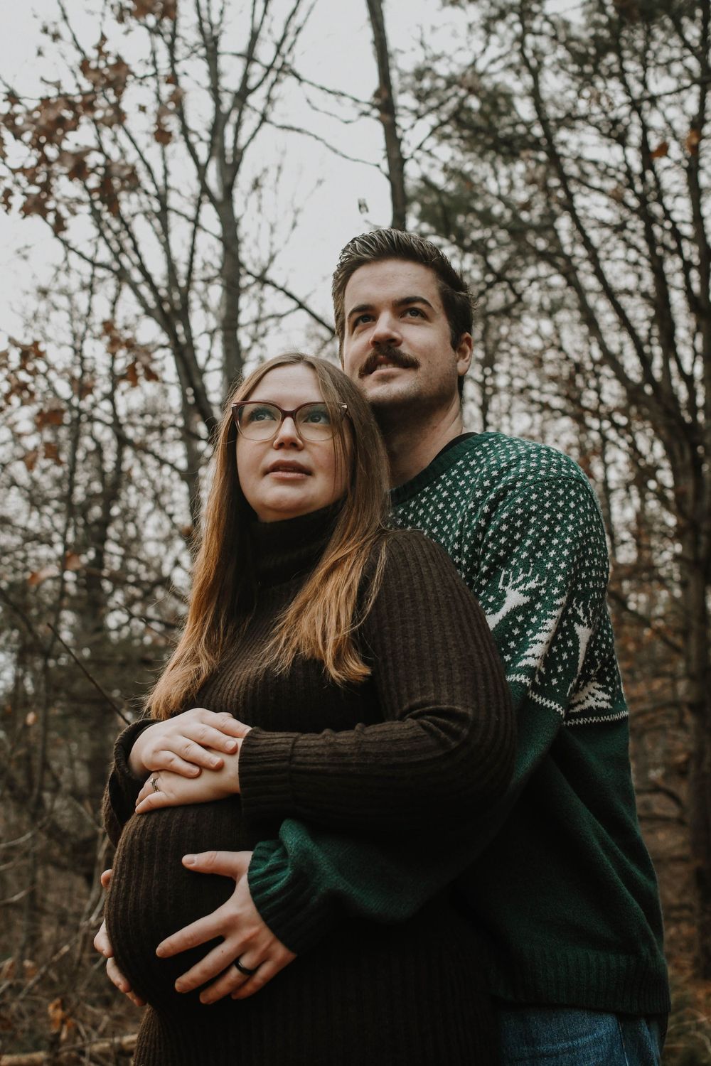Intimate autumn portrait of expecting couple embracing in the woods while wearing cozy sweaters.