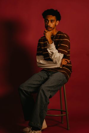 Model sits on stool wearing brown striped sweater and jeans against red background.