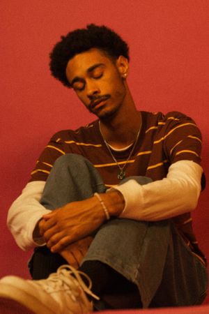 Model in striped sweater sits relaxed with eyes closed against vibrant red background.