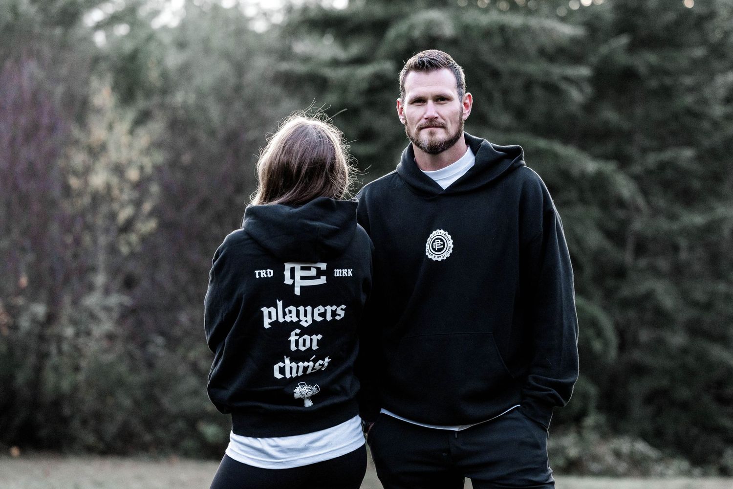 A couple wearing black hoodies stands outdoors, one with a playere for life text design on the back.