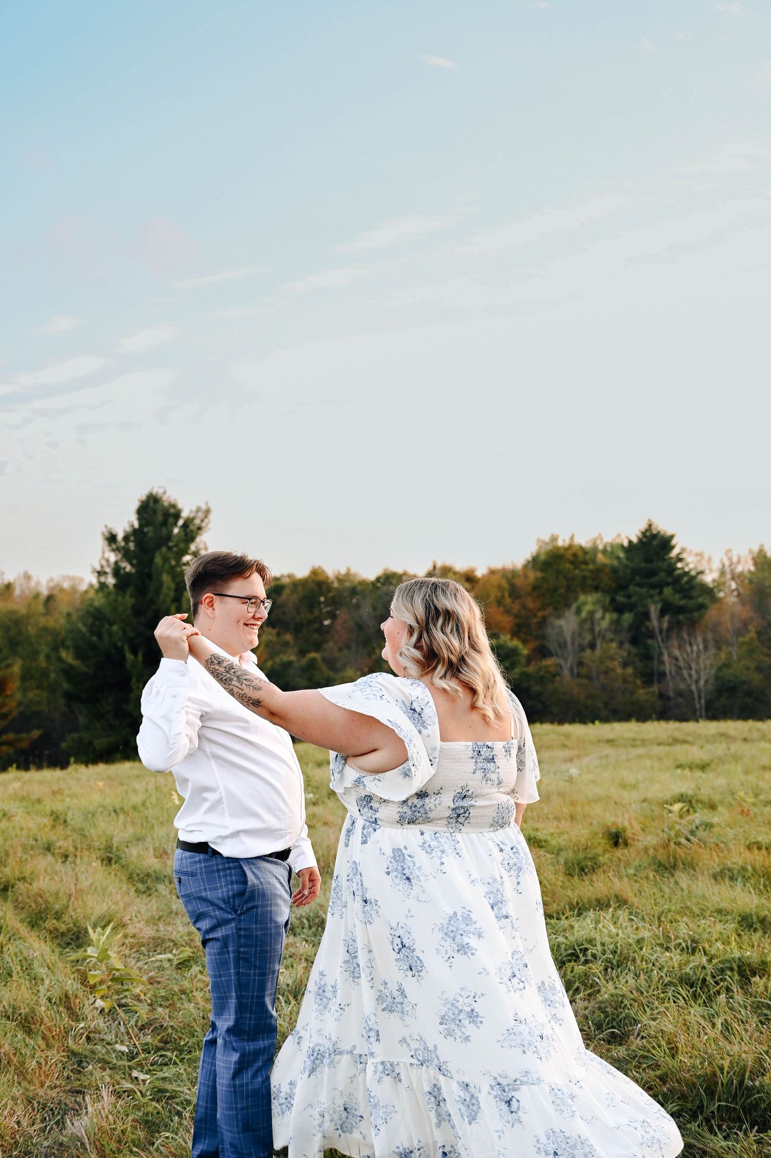 A couple dances together in a field at sunset wearing jeans, a white shirt, and a flowing floral dress.