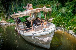 A small white boat with wooden canopy cruises through lush jungle waters at a theme park attraction.