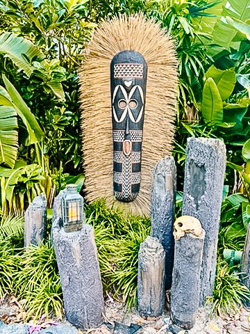 Tiki-style mask decor with grass fringe mounted above stone pillars and lanterns in a tropical garden setting.
