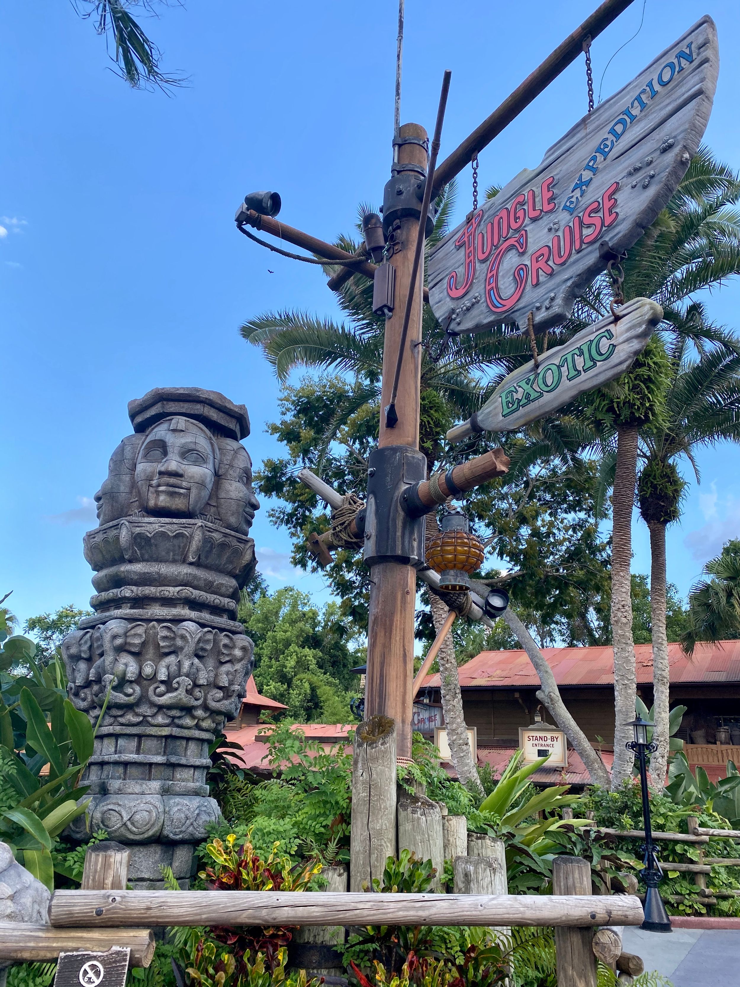 Stone tiki statue and wooden signpost with tropical decor at a theme park entrance with palm trees in background.