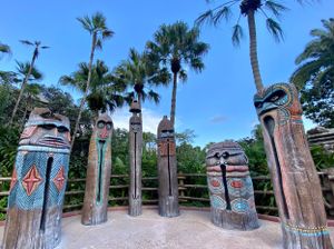 Decorative tiki totems with painted patterns stand in a row under tropical palm trees at a theme park attraction.