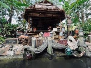 Small rustic wooden stilt house on riverbank with various household items and fishing gear scattered around.