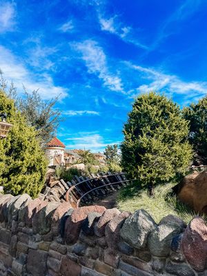 Stone wall and evergreen trees against bright blue sky with wispy clouds on a sunny day.