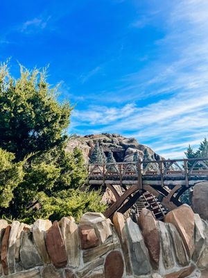 A wooden footbridge crosses over a stone wall formation against a vibrant blue sky with wispy clouds.