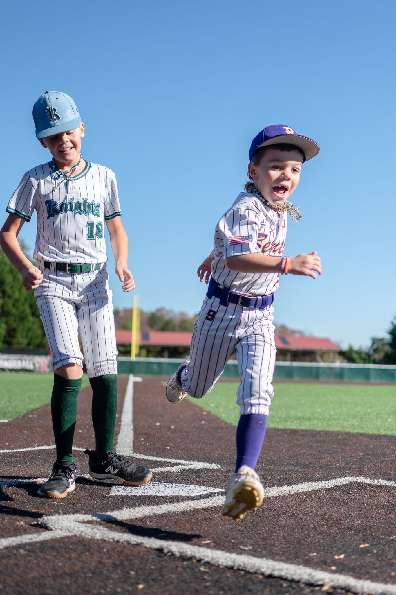Two young baseball players in pinstriped uniforms interact at home plate during a youth baseball game on a sunny day.