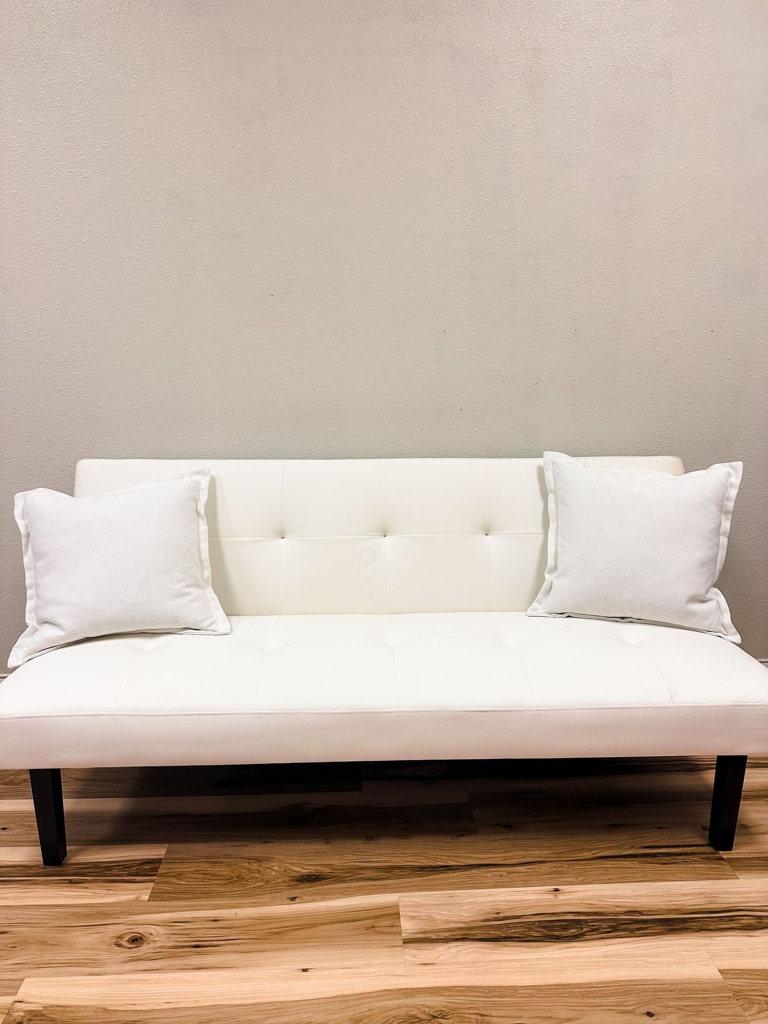Modern white sofa with tufted backrest and decorative pillows sits on hardwood flooring against beige wall.