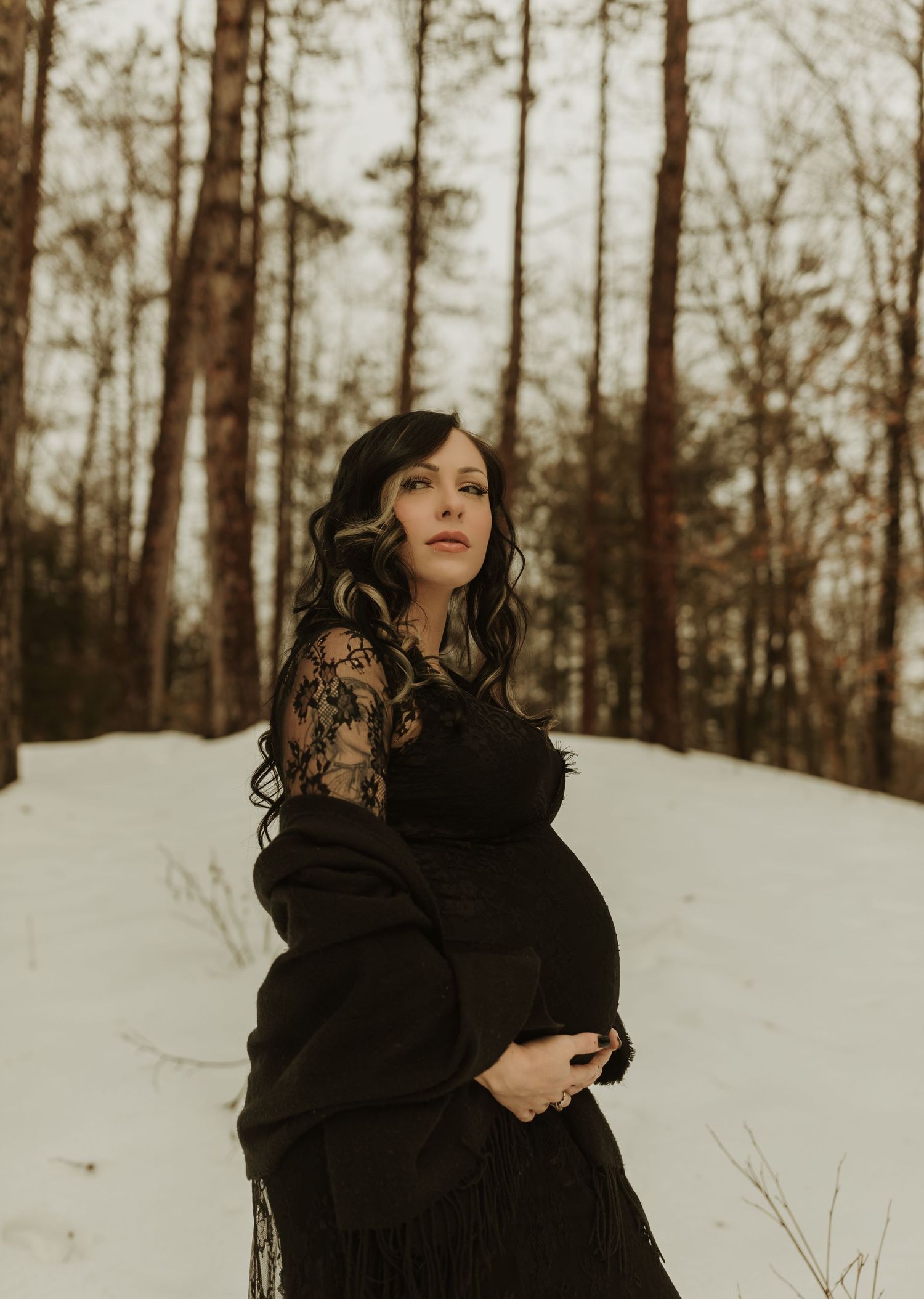 A dramatic maternity portrait in a snowy forest with woman wearing a black lace dress.