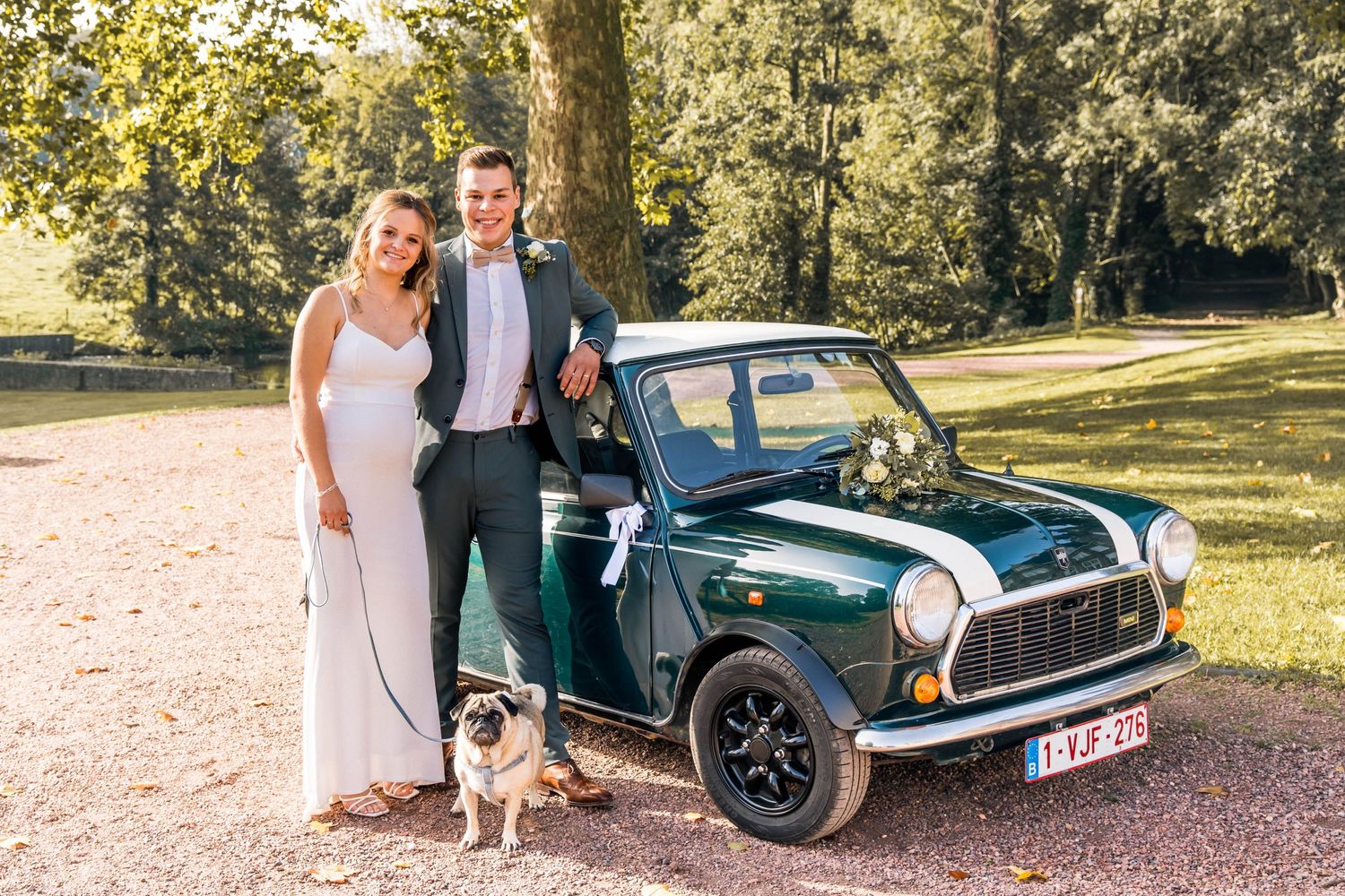 A couple poses with their dog next to a classic green Mini Cooper in a sunny park.