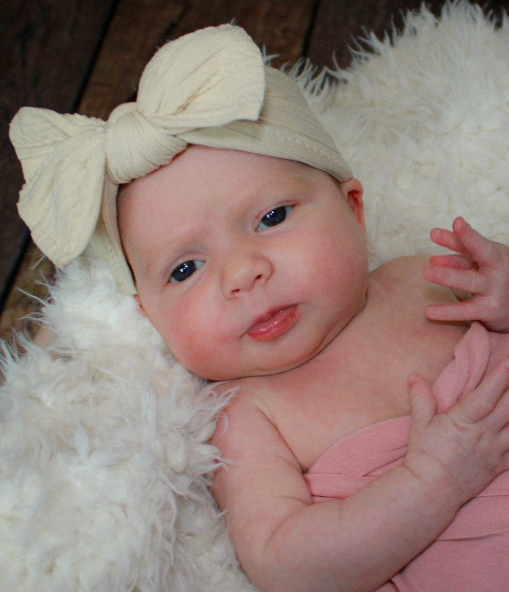 Baby wearing a cream bow headband lying on soft white fur.