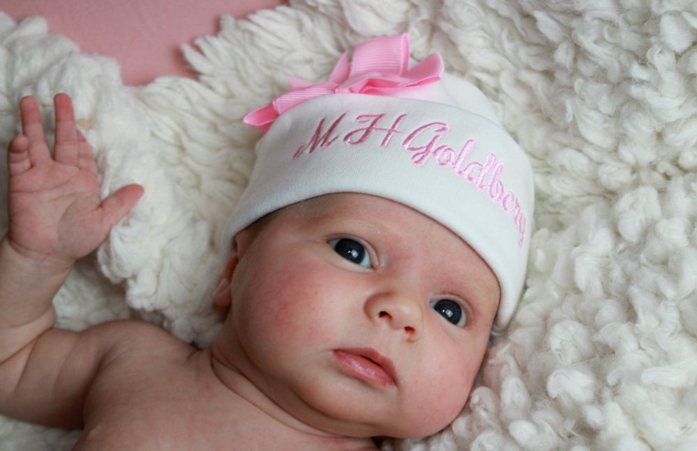 Baby on white fur wearing a personalized white hat with pink lettering.