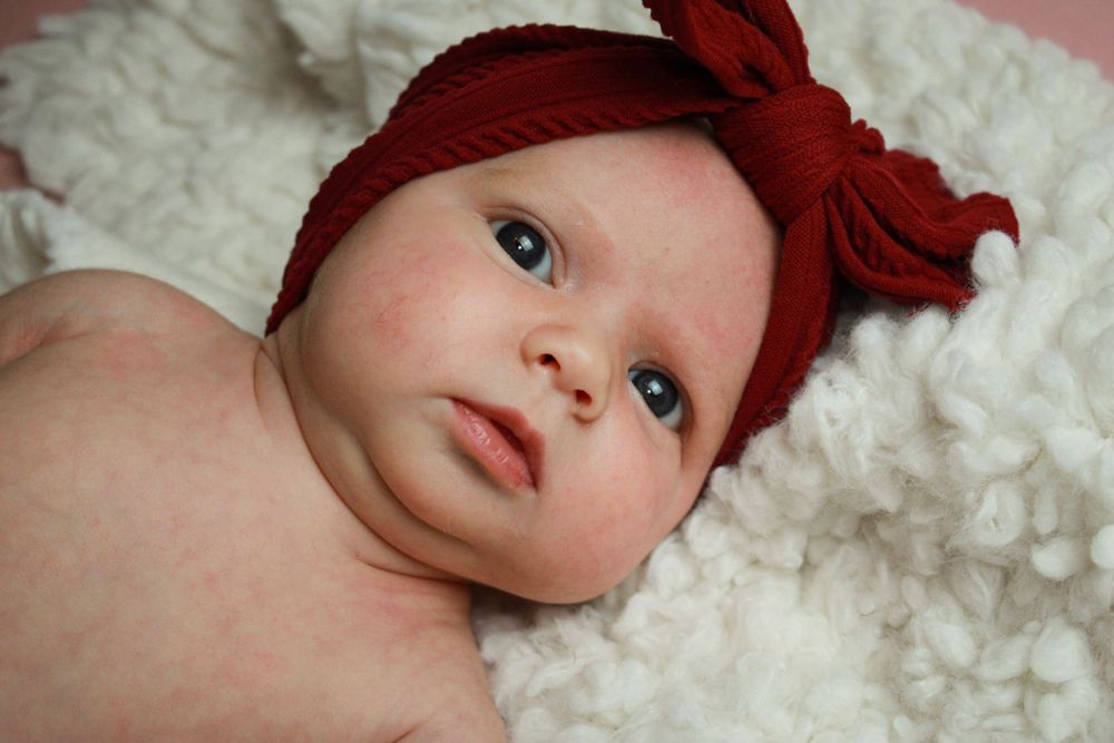 Baby in a red headband with bow resting on white textured blanket.