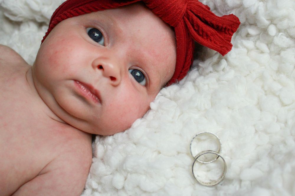 Baby with red bow headband lying next to wedding rings on white fur.