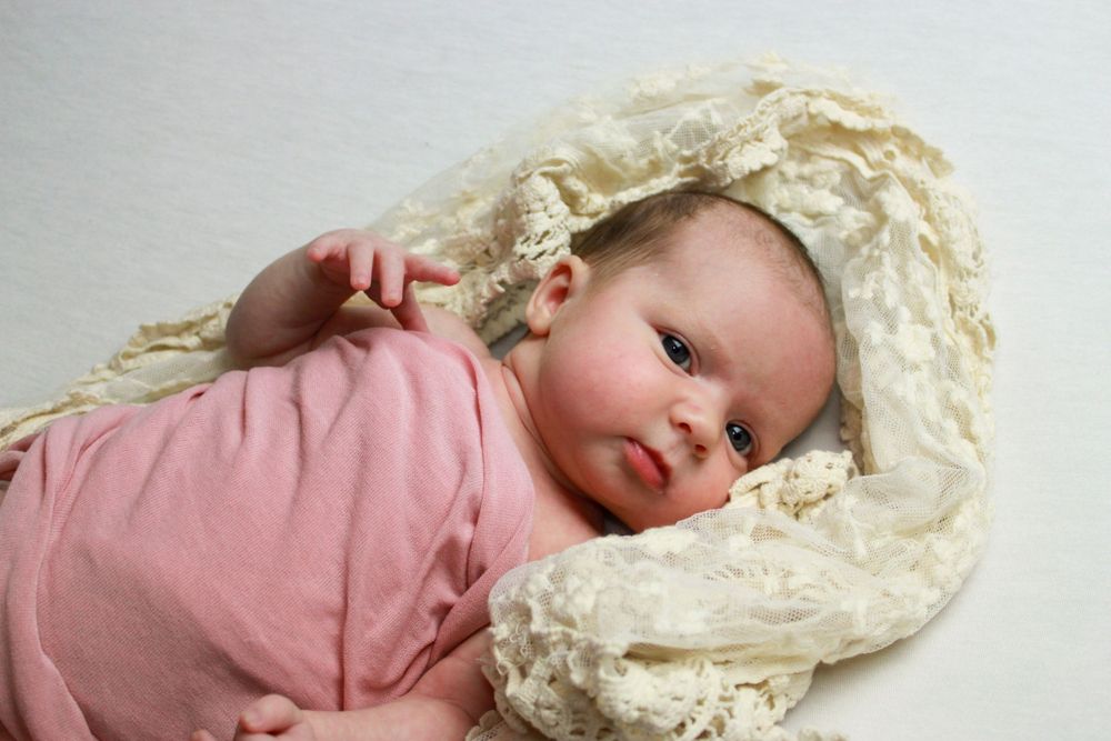 Newborn baby wearing pink clothing laying on cream-colored lace fabric.