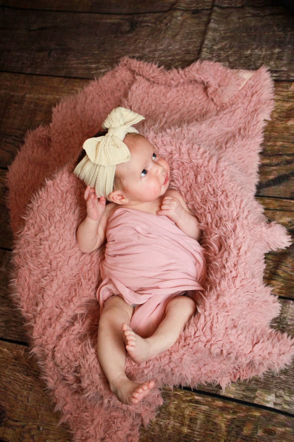 Baby in pink wrap lying on fluffy pink rug with white bow headband.