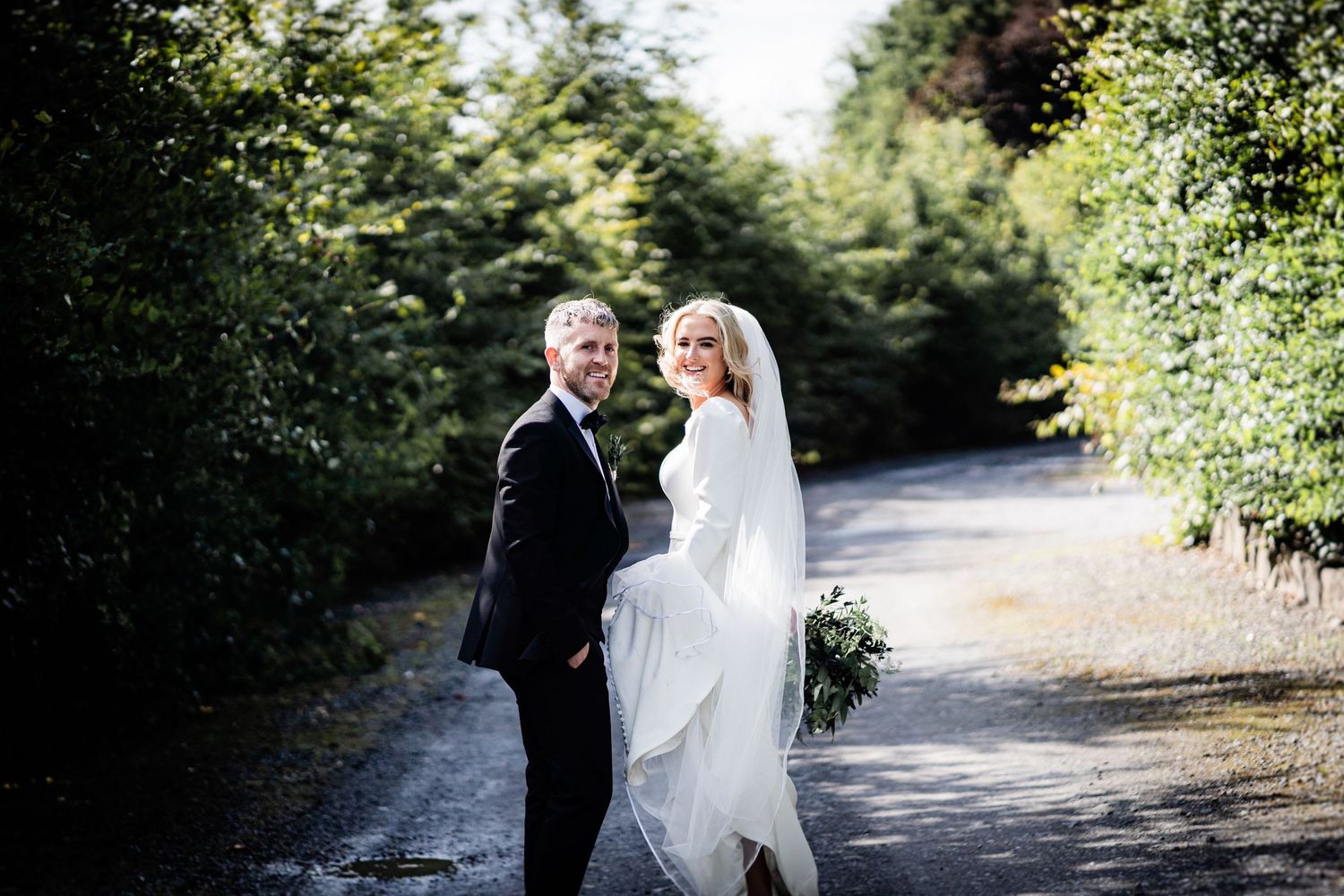 A couple in wedding attire standing on a tree-lined path outdoors.