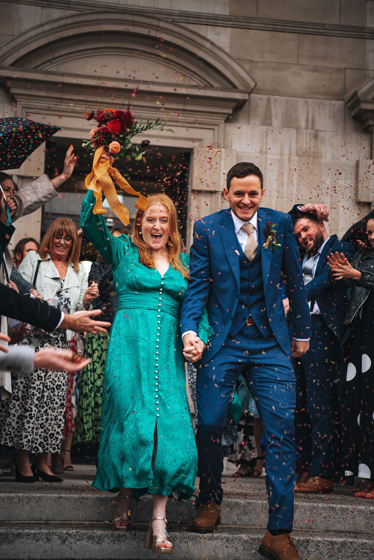 bride in turquoise holding bouquet above her head holds grooms hand as they walk through confetti tunnel