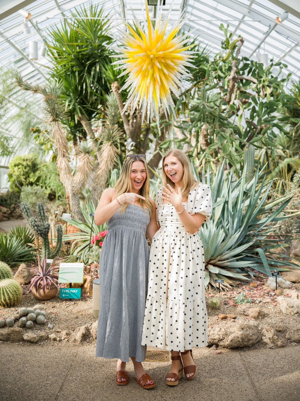 Two women in summer dresses laughing in a greenhouse with cacti and a yellow glass sculpture.