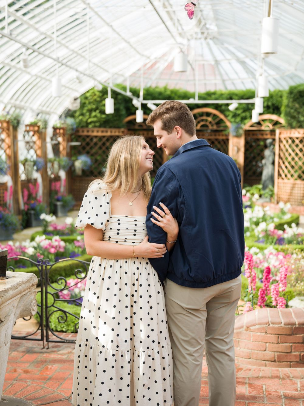 A couple embraces and smiles at each other inside a beautiful flower-filled greenhouse, woman wearing a polka dot dress.
