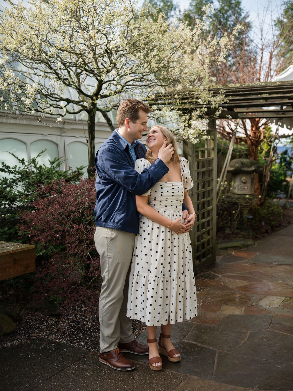 Couple embracing in a garden, woman in polka dot dress, man in blue jacket, blooming trees in background.