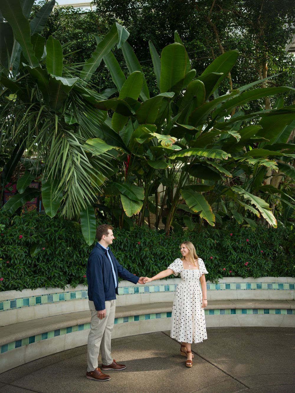 A couple holds hands and dances surrounded by lush tropical plants in a botanical garden setting.