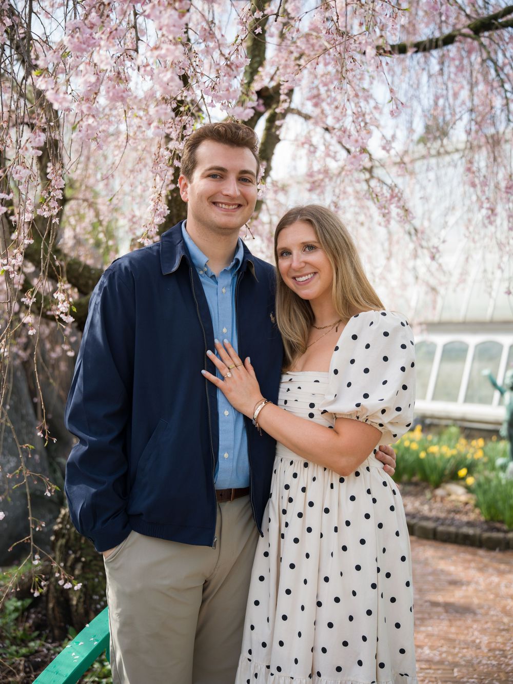 Couple smiling under blooming pink cherry blossom tree, woman in polka dot dress showing engagement ring.