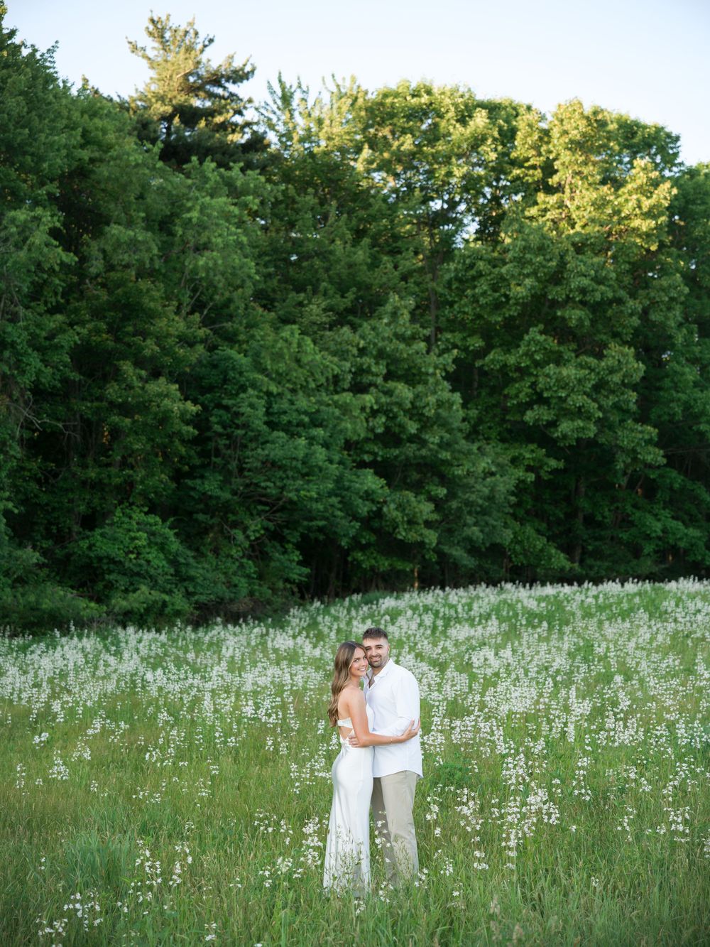 Couple embracing in a lush green meadow filled with white wildflowers, surrounded by tall trees at golden hour.