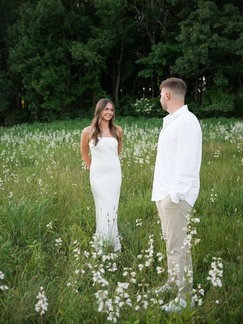 A couple in white attire stands in a wildflower meadow, sharing a romantic moment with lush green trees behind them.