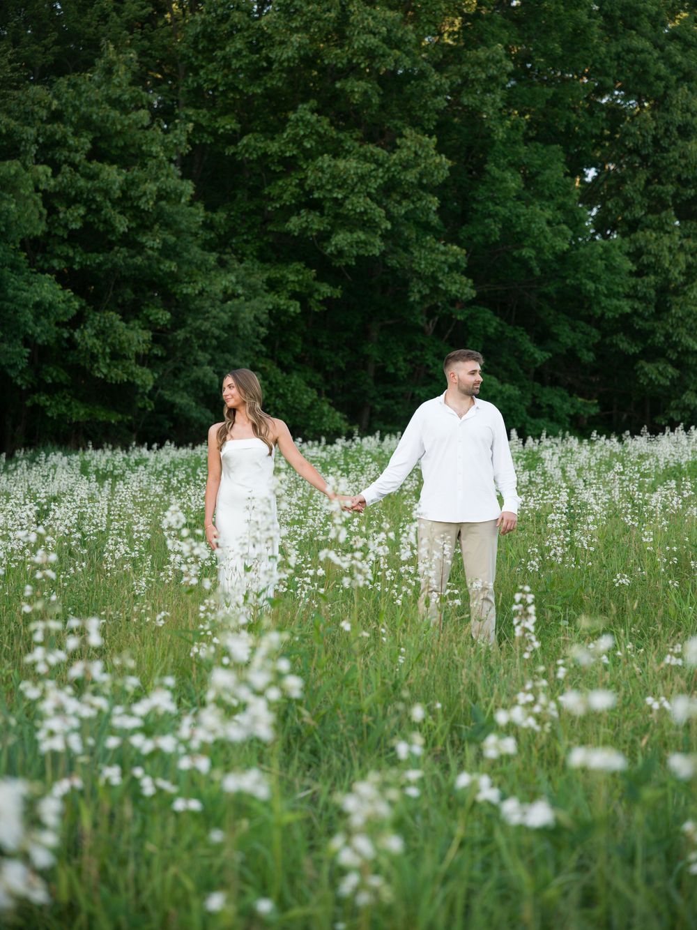 Couple holding hands in a white wildflower meadow with lush green trees, wearing white and beige outfits.