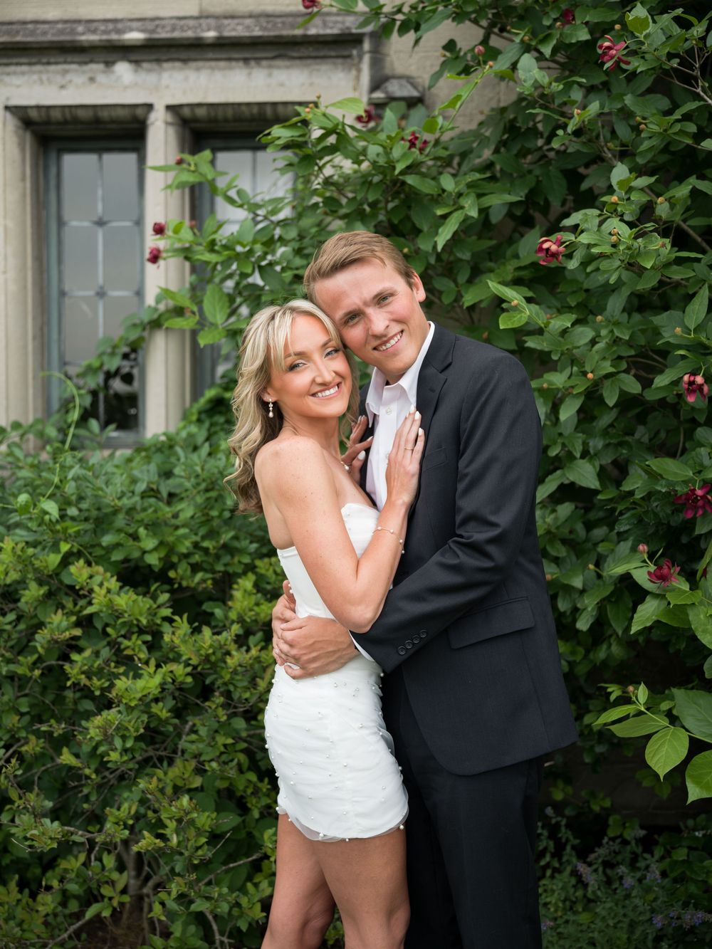 Couple in wedding attire embracing outdoors, woman in white mini dress and man in black suit, posing before lush greenery.