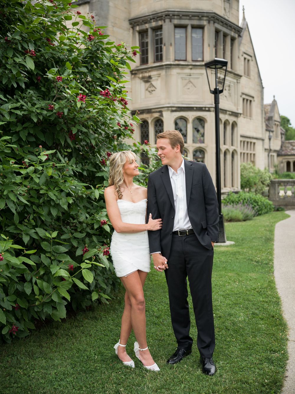 Couple in wedding attire holding hands beside lush greenery near a historic stone manor building.