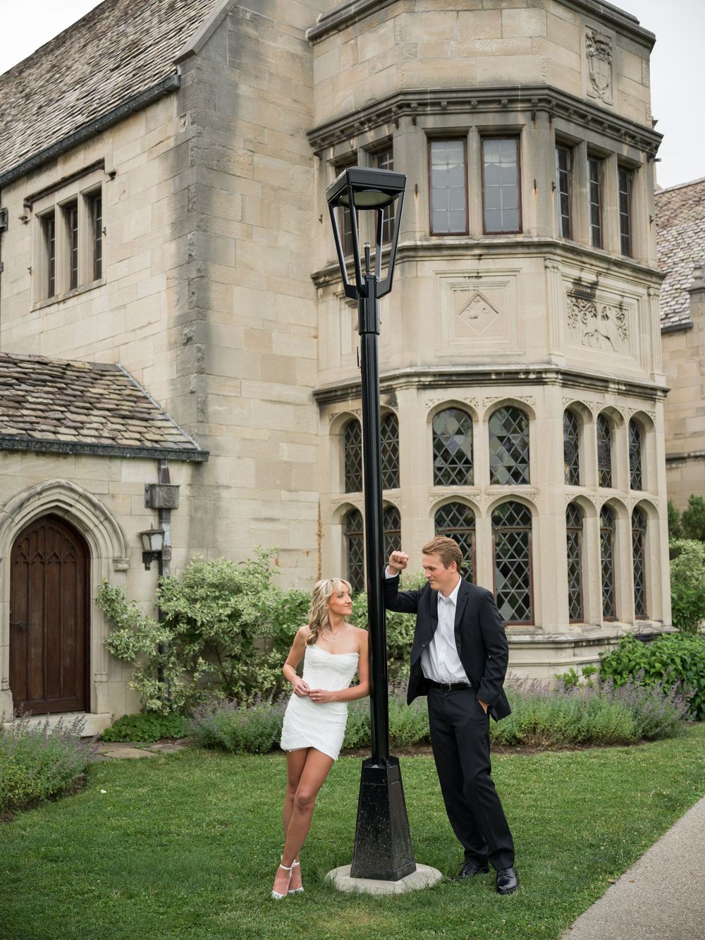 Couple posing by a vintage lamppost outside an elegant stone manor, woman in white dress, man in black suit.