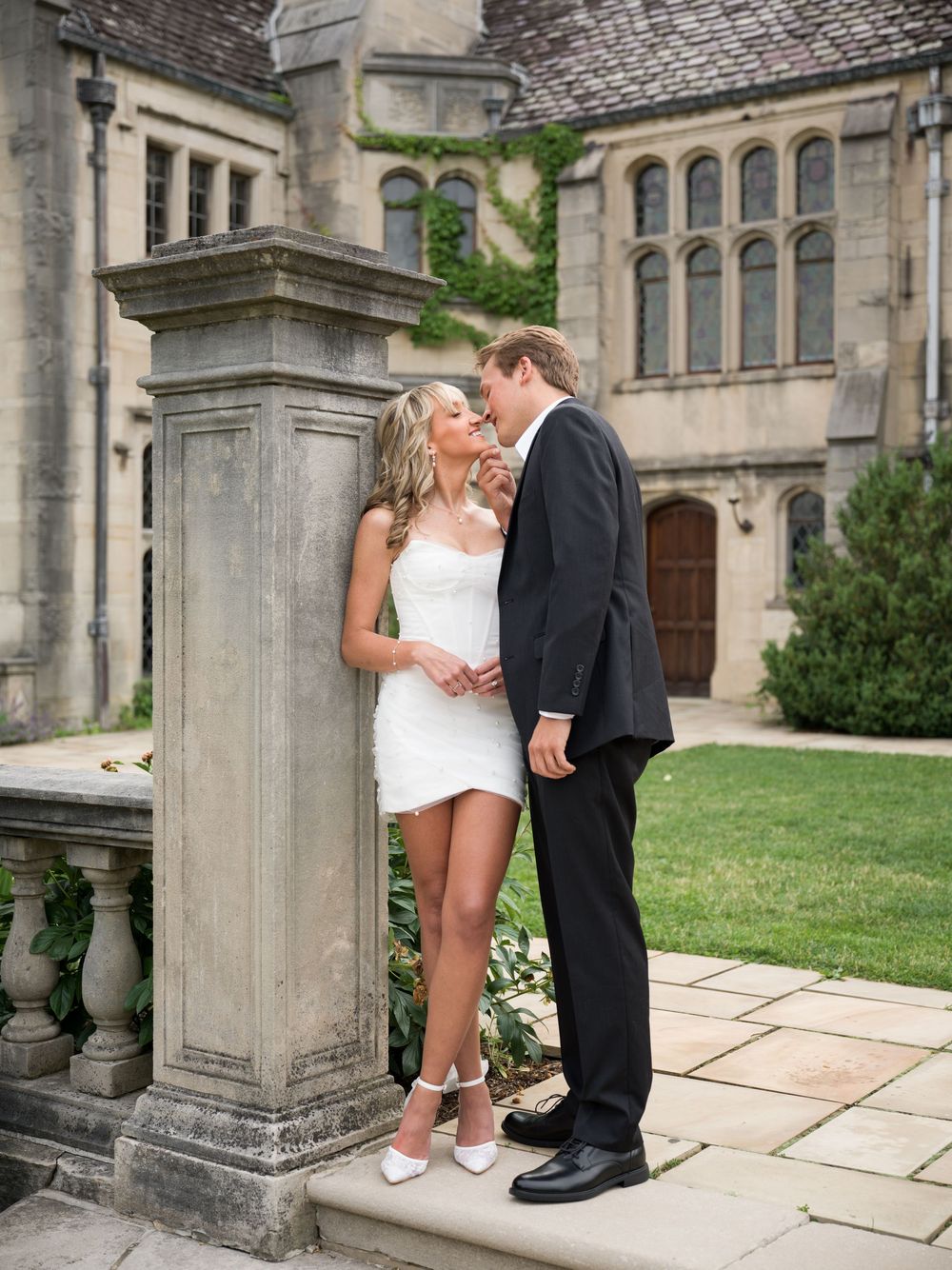 Bride in short white dress and groom in black suit share a kiss by a stone pillar at an elegant manor estate.