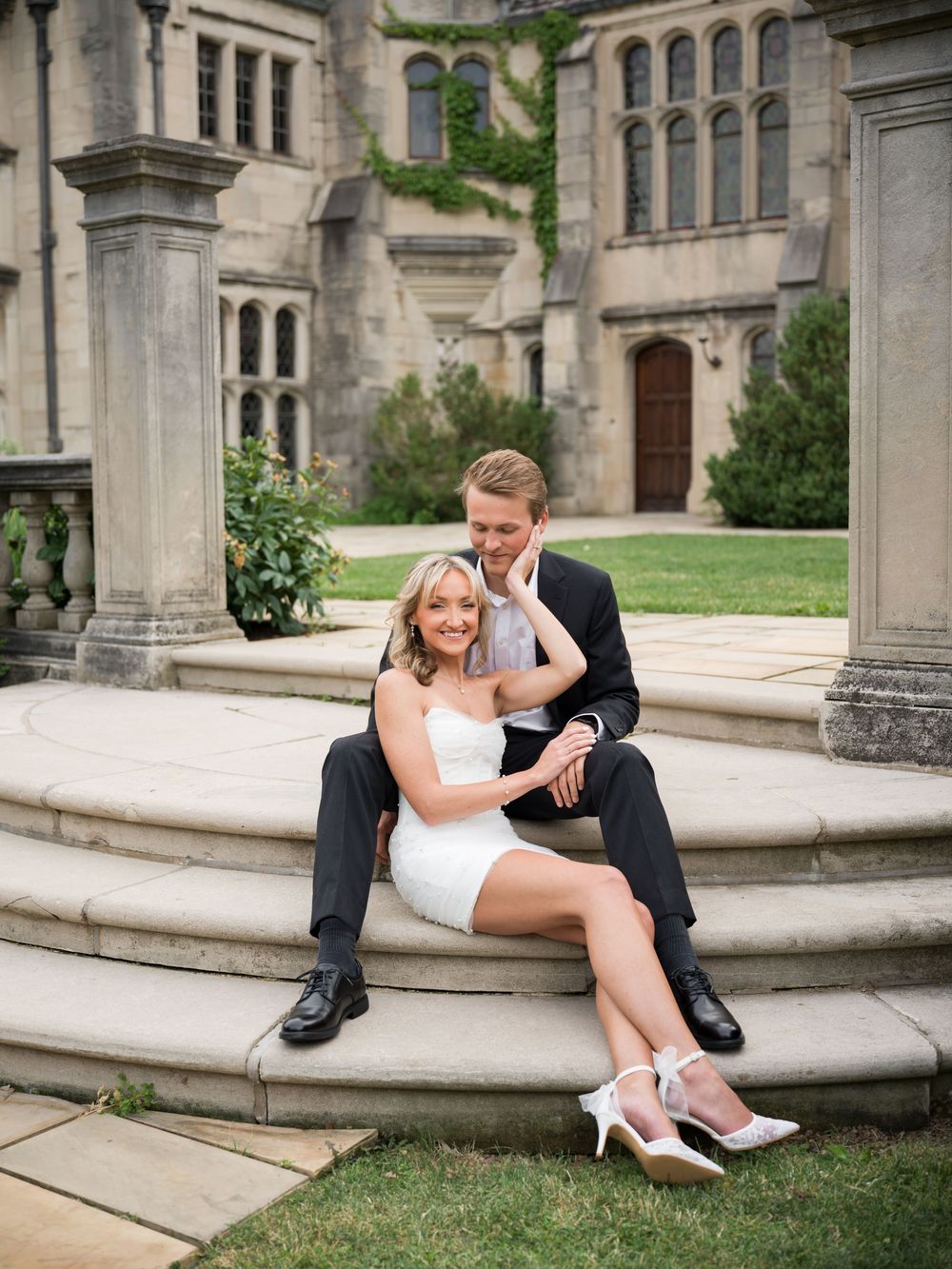 Couple in wedding attire sitting on stone steps outside a grand Gothic manor, bride in white mini dress, groom in black suit.