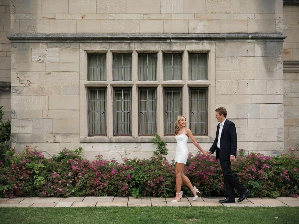 Couple in wedding attire holding hands walking past flowering bushes beside a stone building.