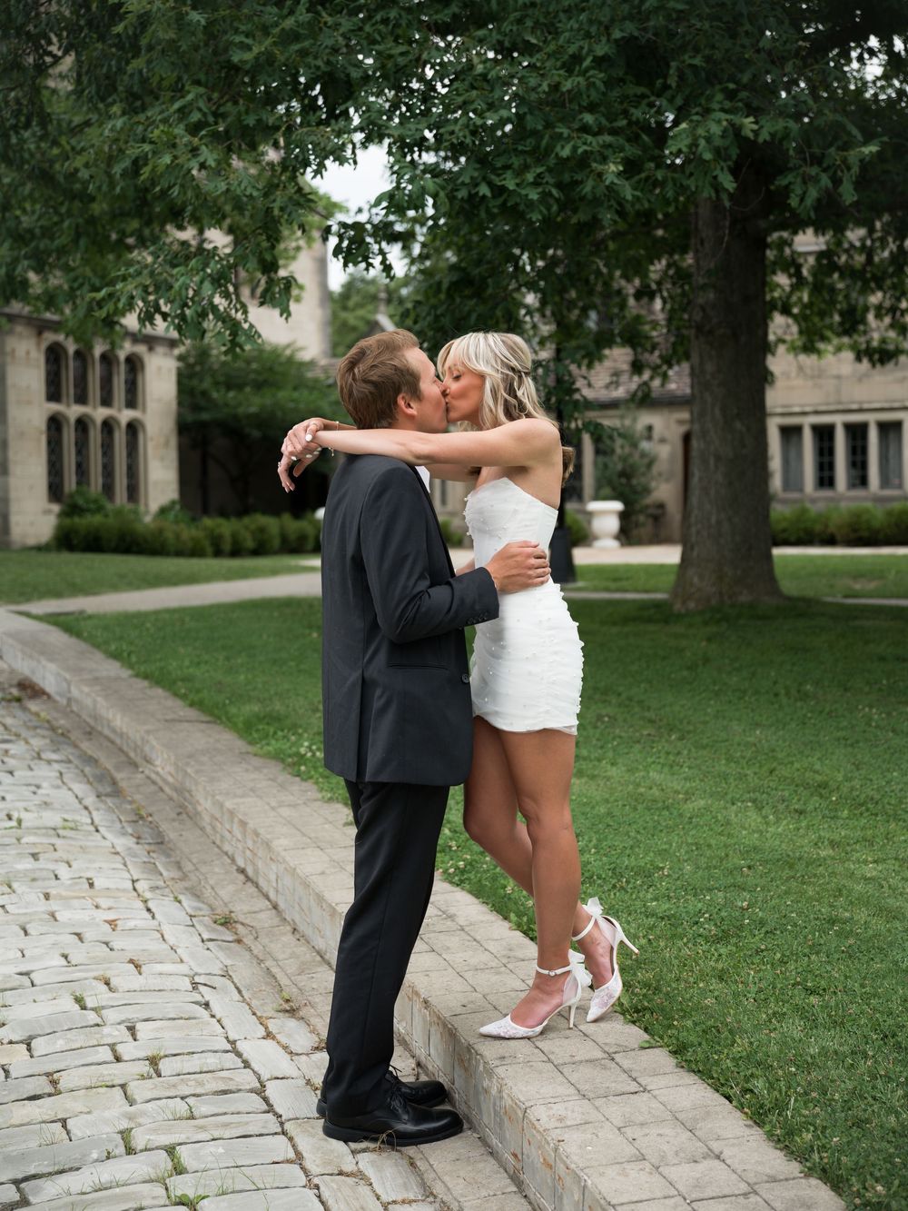 Couple sharing a romantic kiss outdoors, bride in short white dress and heels, groom in black suit, on a stone path.