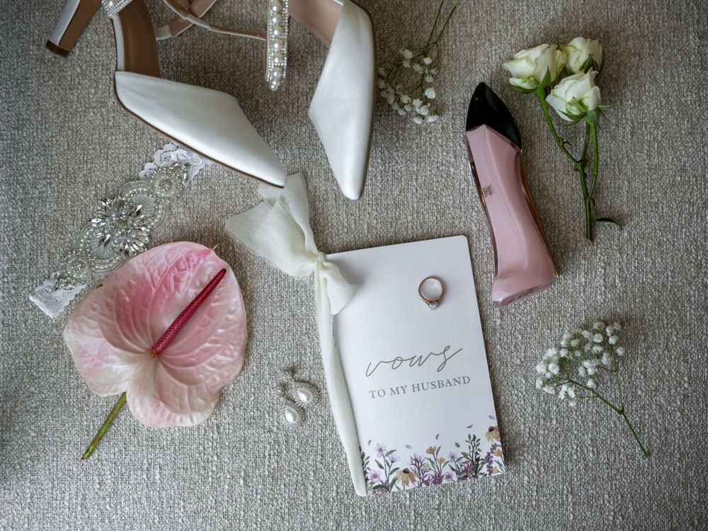 Wedding details flatlay with white shoes, pink flowers, and vow book on grey linen background.