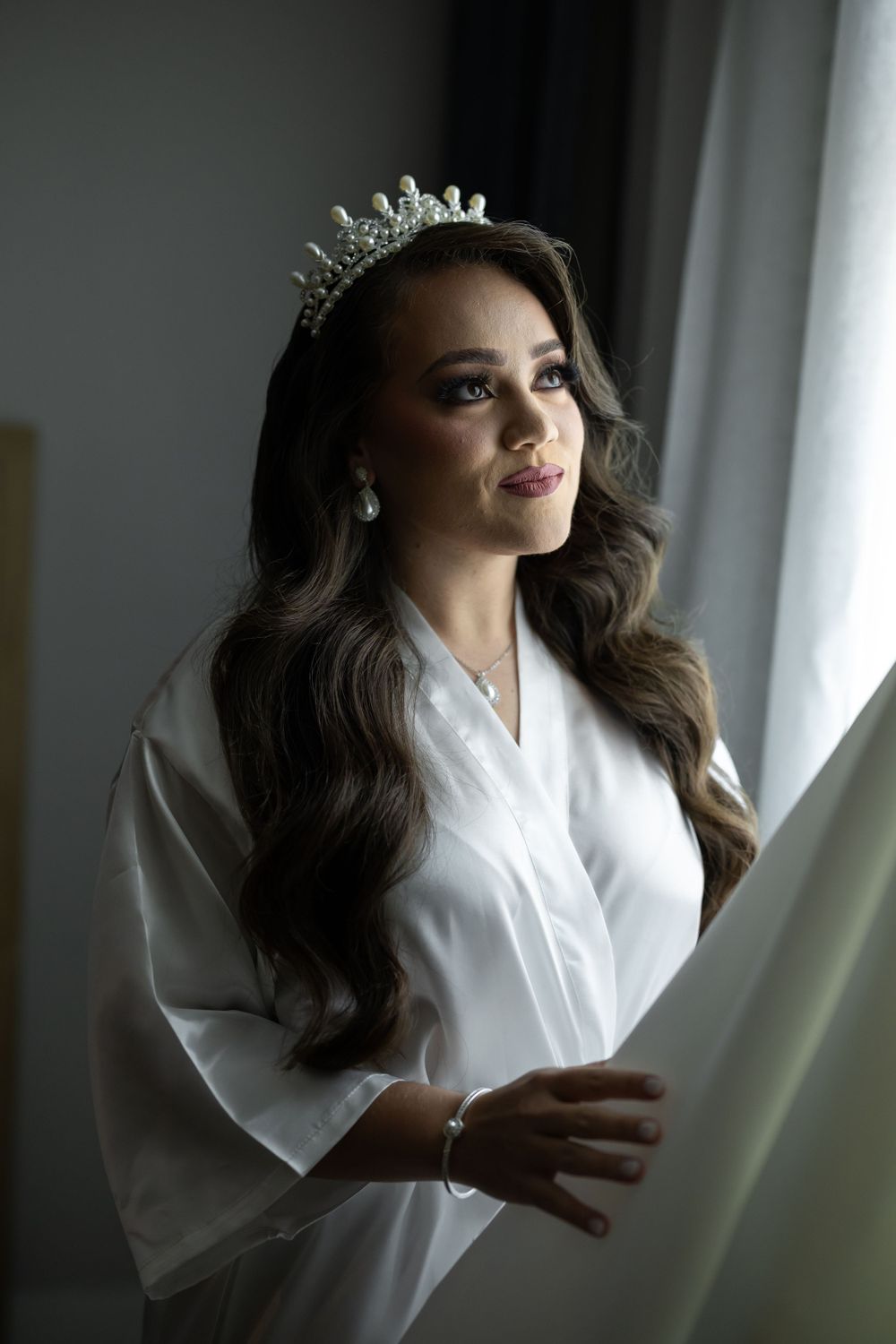 Woman in white robe wearing crystal tiara and glamorous makeup by window.