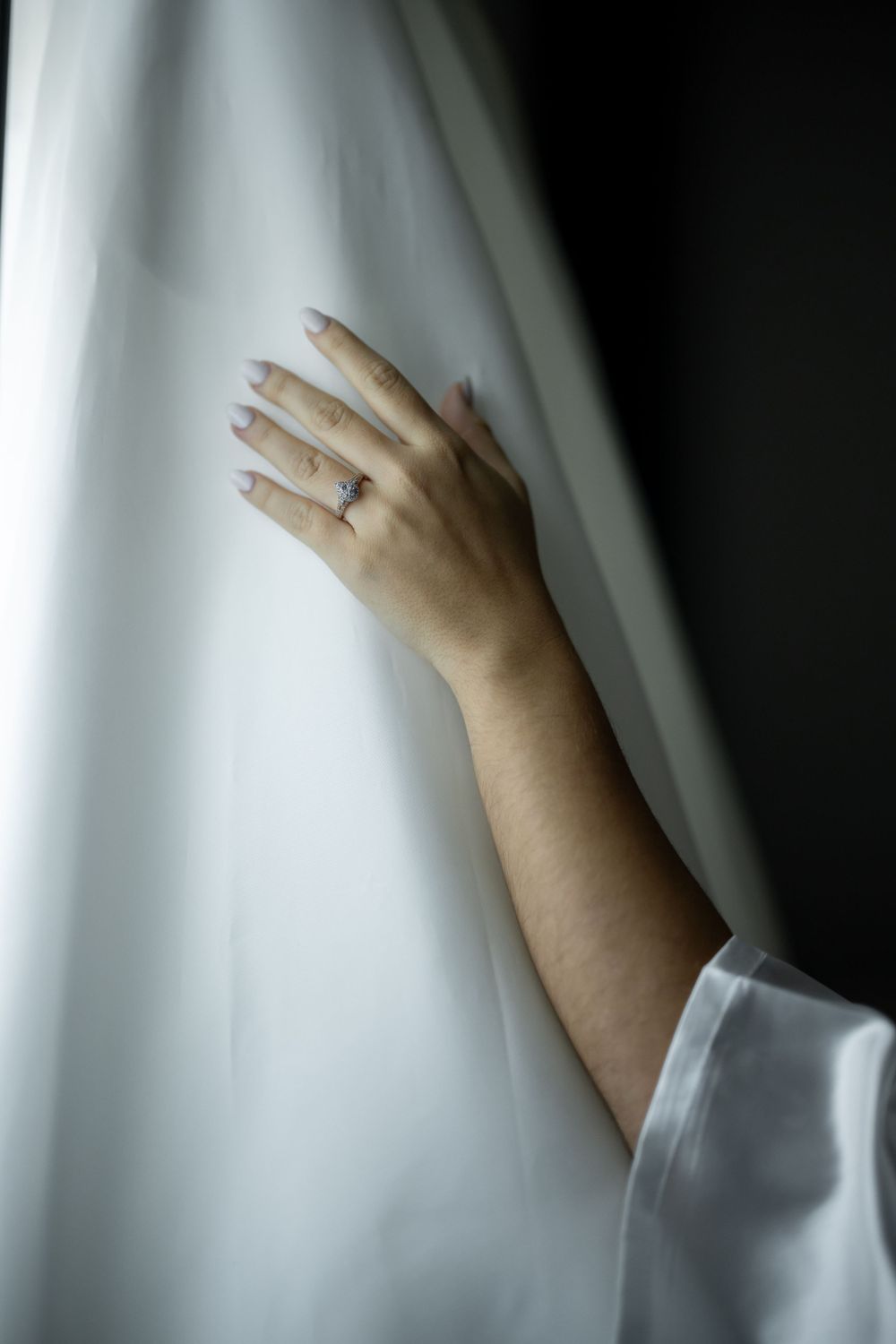 Hand with engagement ring gently touching white fabric in moody lighting.