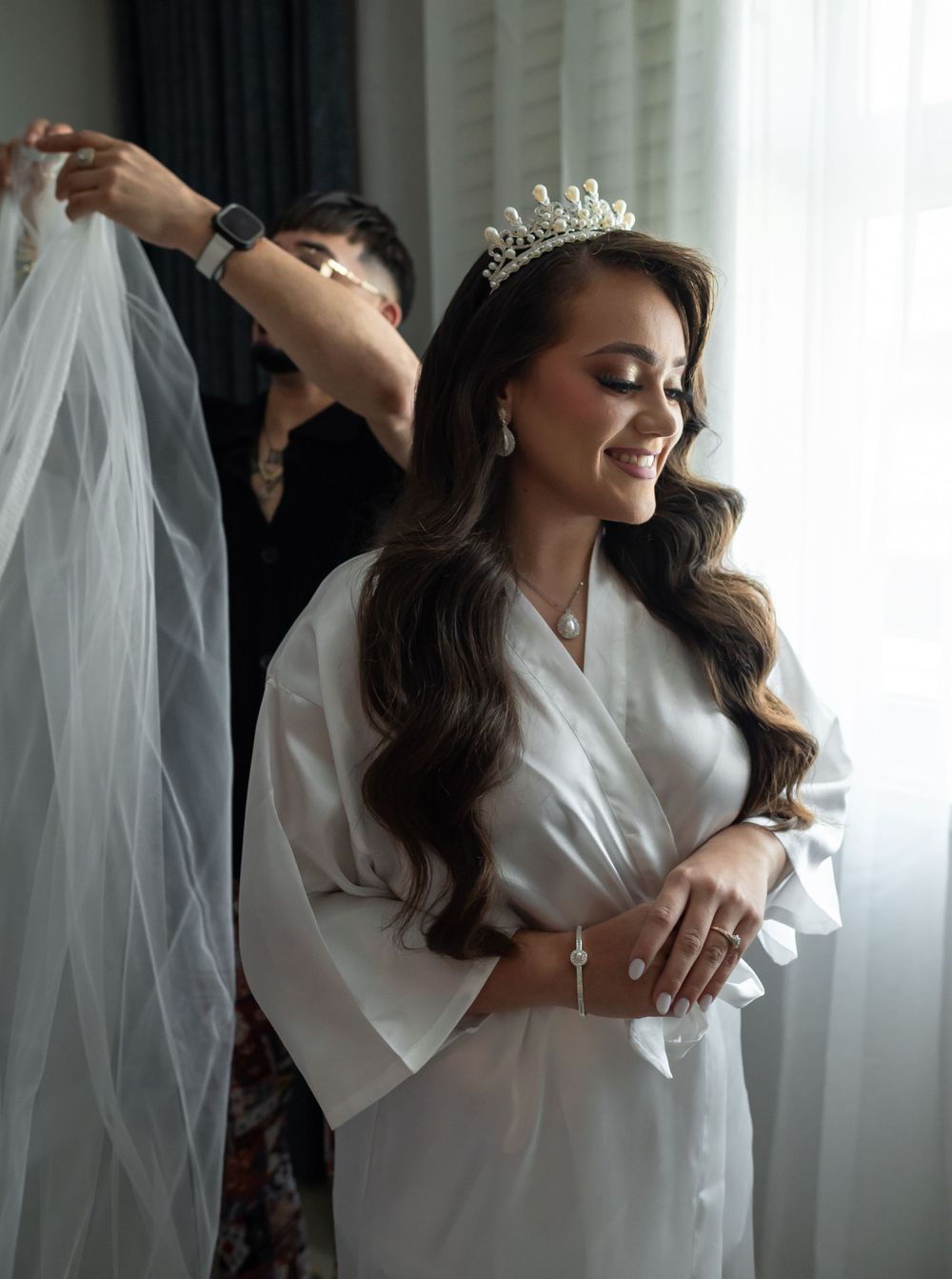 Bridal preparation scene with helper adjusting veil as bride smiles in white robe.