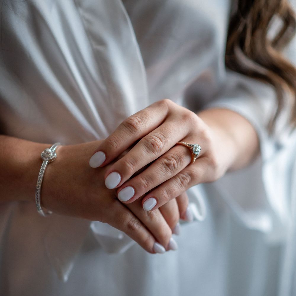 Manicured hands with engagement ring and bracelet folded gracefully against white robe.