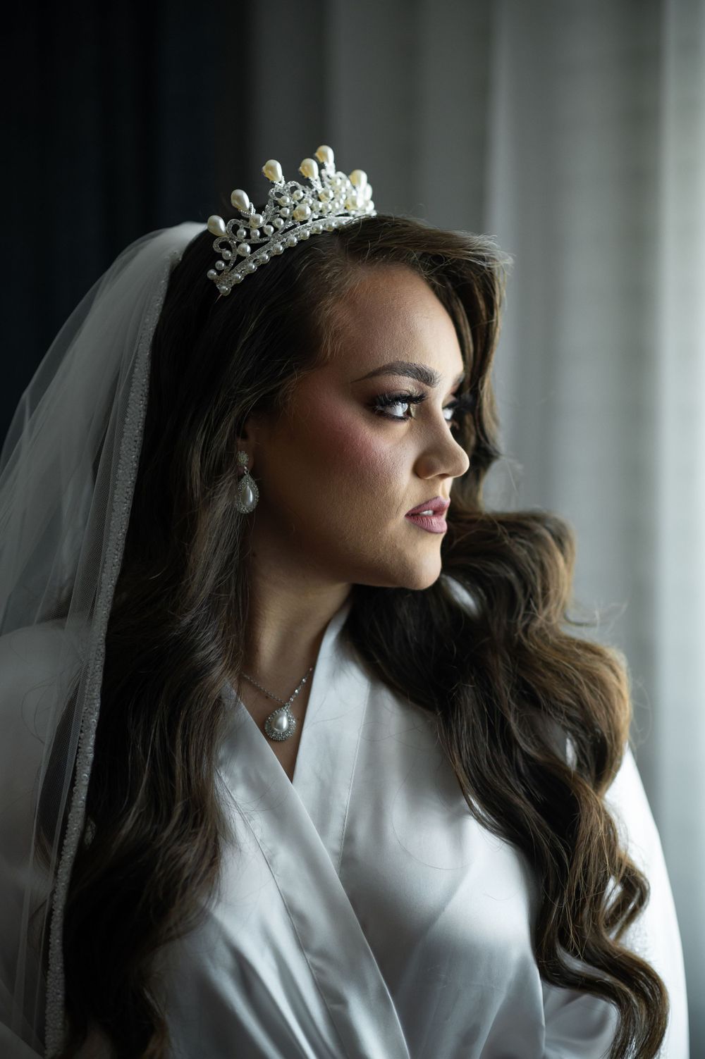 Bride in white robe wearing crystal tiara and veil in dramatic window lighting.