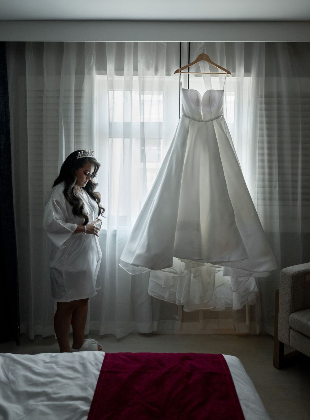 A wedding dress hangs elegantly in front of sheer curtains as morning light streams in.