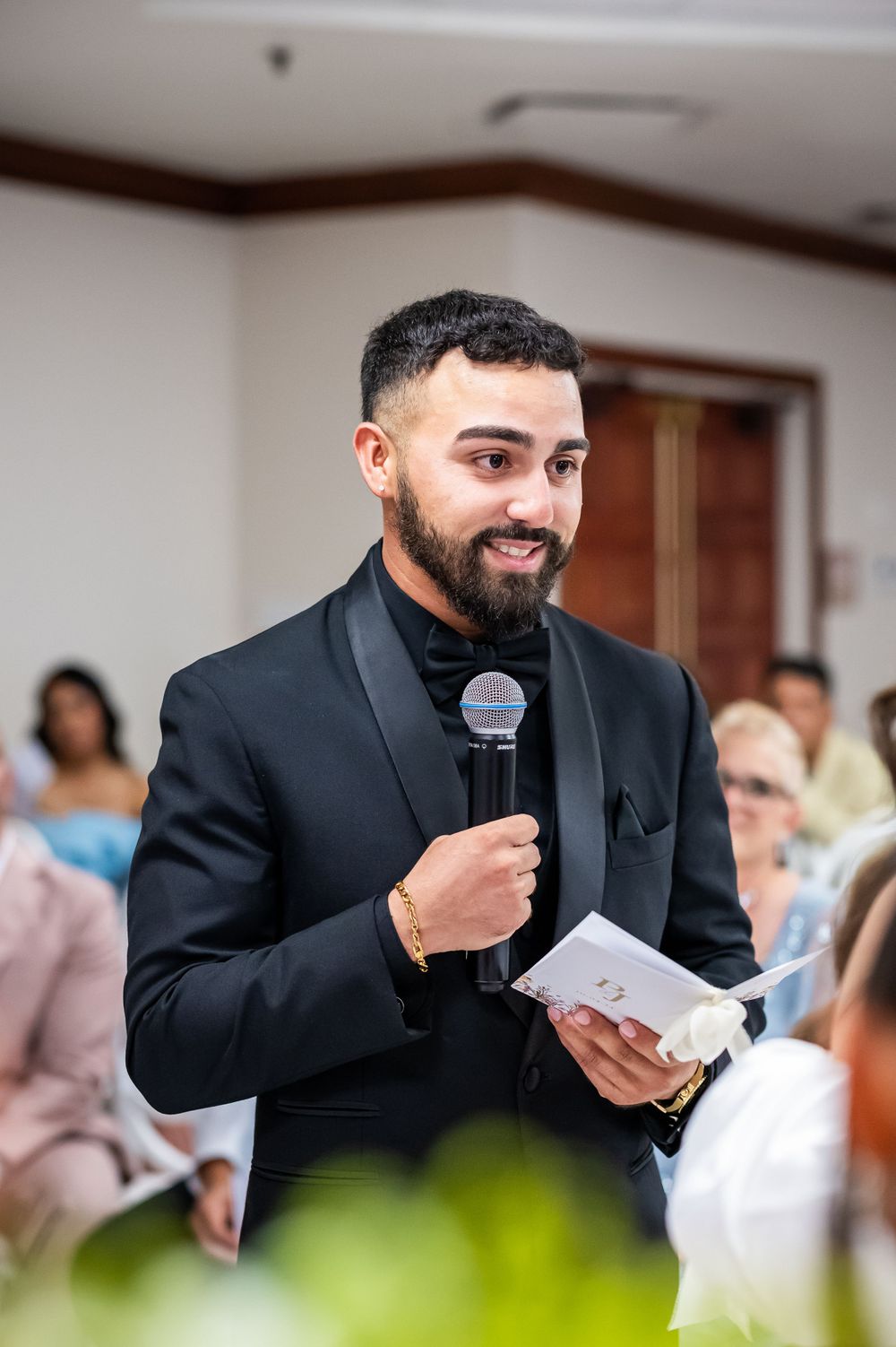 Person in black suit giving animated speech at wedding reception with microphone in hand.